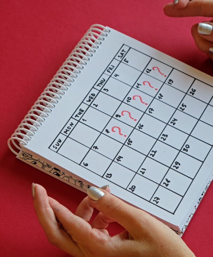 A woman crosses her fingers while looking at a calendar where five days are marked with a red question mark